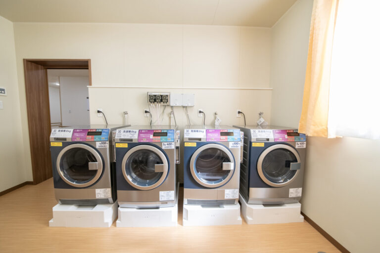 共用の洗濯室。複数の大型洗濯機が整列し、入居者が利用できる設備。